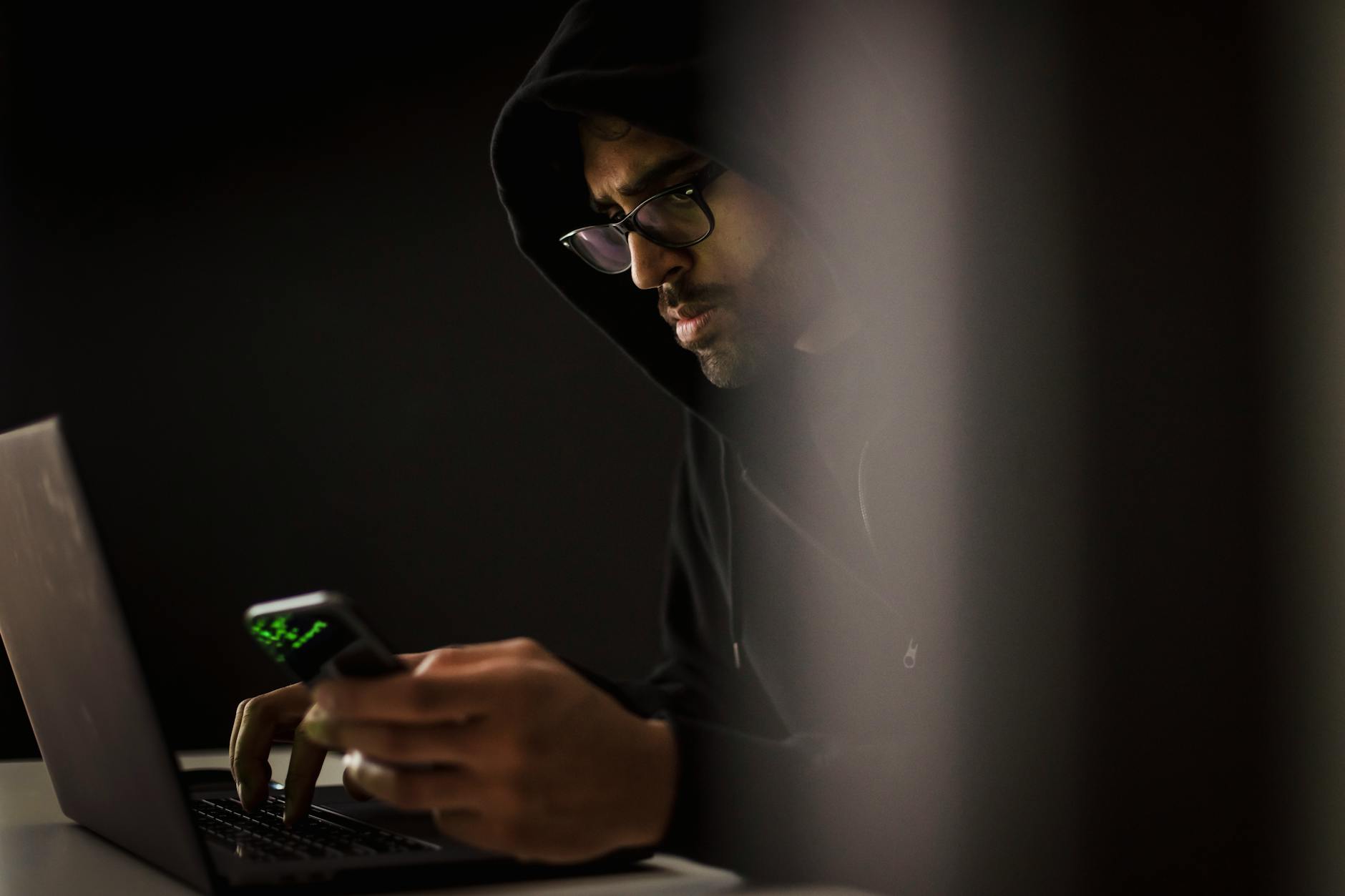 Brodaty mężczyzna w kapturze korzysta z telefonu i laptopa w ciemnym pokoju